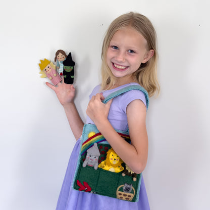 Young girl in purple dress holding a felt puppet bag for kids with wizard of oz themed characters of Dorothy, the Tin Man, The Cowardly Lion, Glinda the Good Witch, The Wicked Witch of the West, and the Scarecrow.