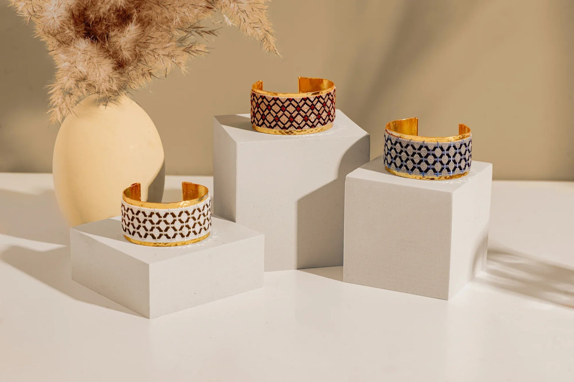 Gold plated cuff with blue embroidery artisan design on white display block with other similar bracelets on other white blocks.