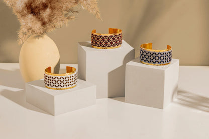 Gold plated cuff with blue embroidery artisan design on white display block with other similar bracelets on other white blocks.