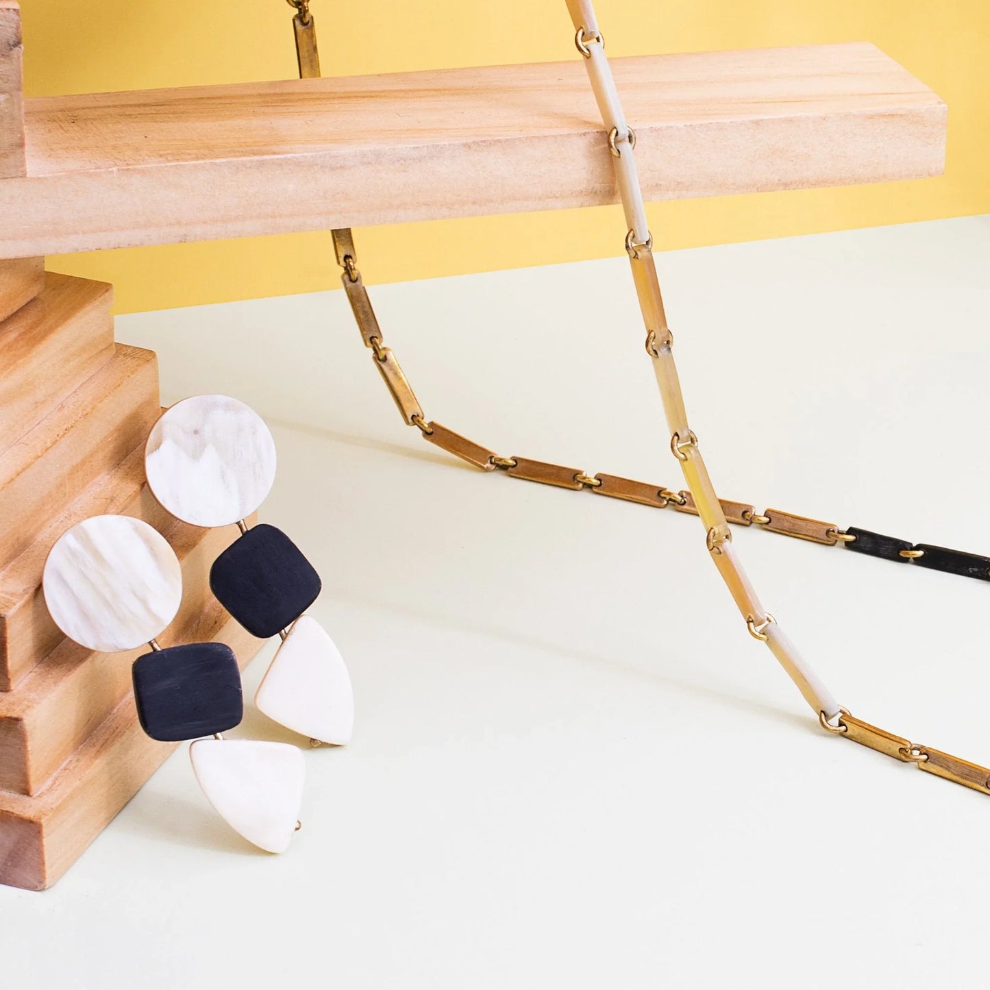 White, gold, and black link necklace hanging from a wood rod next to white and black geometric earrings.