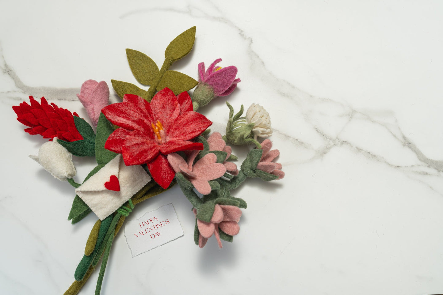 Bouquet of pink, red, and white felt flowers and green leaves on marble surface.