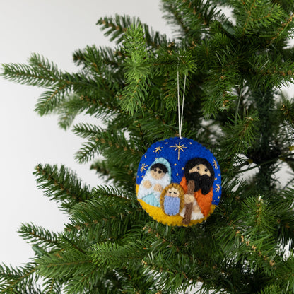 Felt ornament in a circle shape showing the nativity scene of Mary, Joseph, and baby Jesus underneath the night sky hanging on a tree.