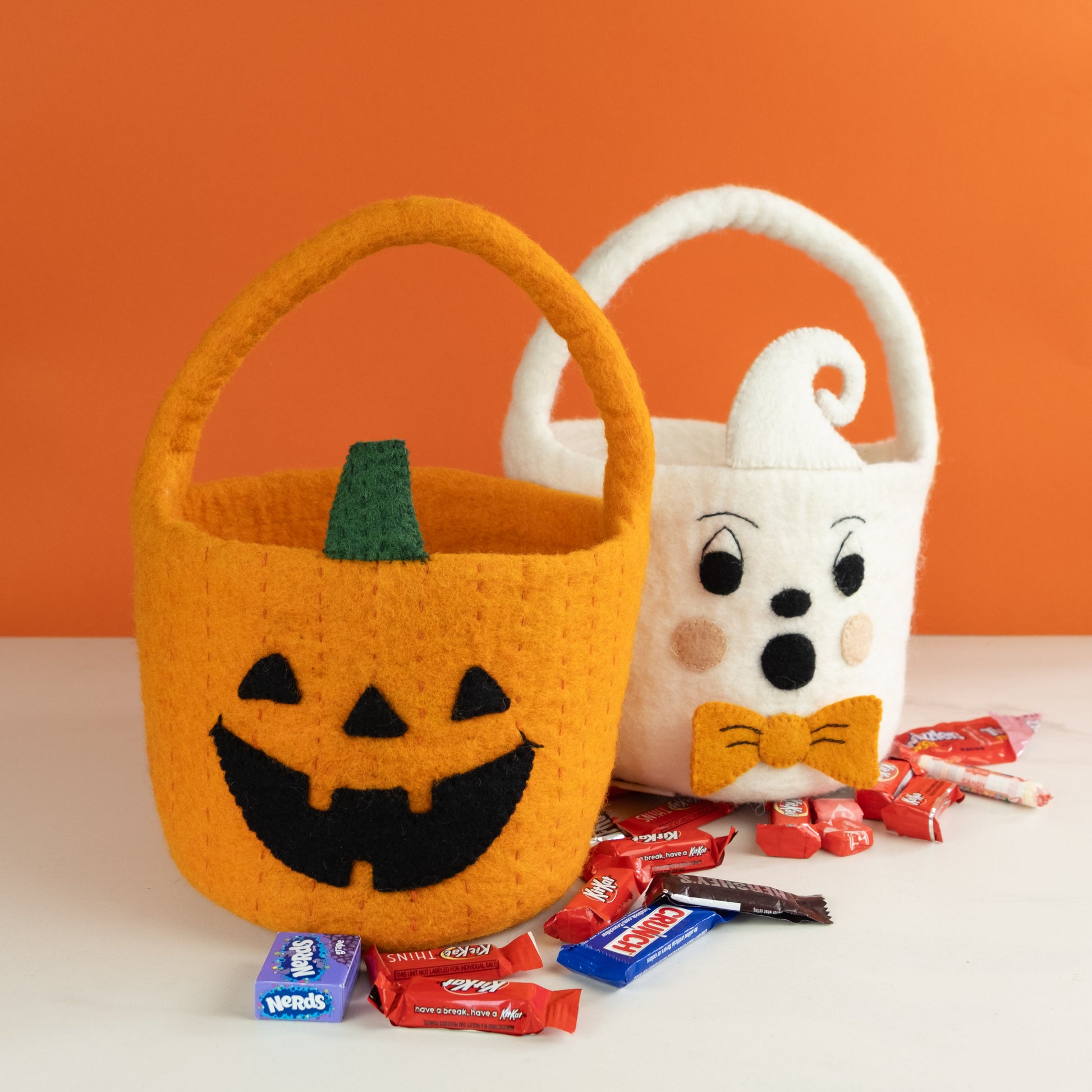 Orange felt jack-o-lantern trick-or-treat bucket and a white felt ghost trick-or-treat bucket with orange bow tie and candy below them.