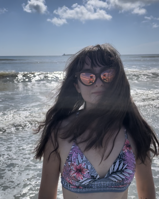 Woman with brown hair on the beach wearing dark brown bamboo cat eye sunglasses with pink mirrored lenses and a pink tropical bikini top.