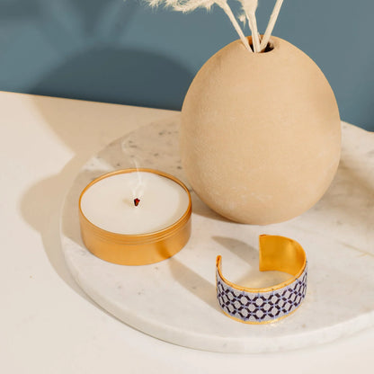 Gold plated cuff bracelet with blue embroidery artisan design on a white tray with candle and diffuser.