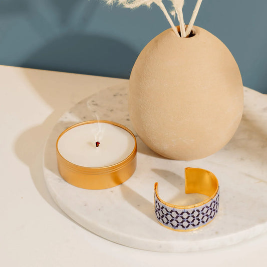 Gold plated cuff bracelet with blue embroidery artisan design on a white tray with candle and diffuser.