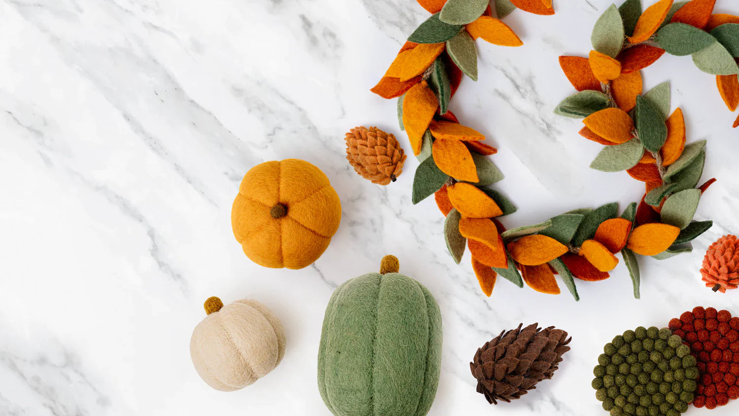 Three felt pumpkins, one large orange pumpkin, one medium oblong green one, and one small tan one spread out on white counter with pinecones and fall leaf garland.
