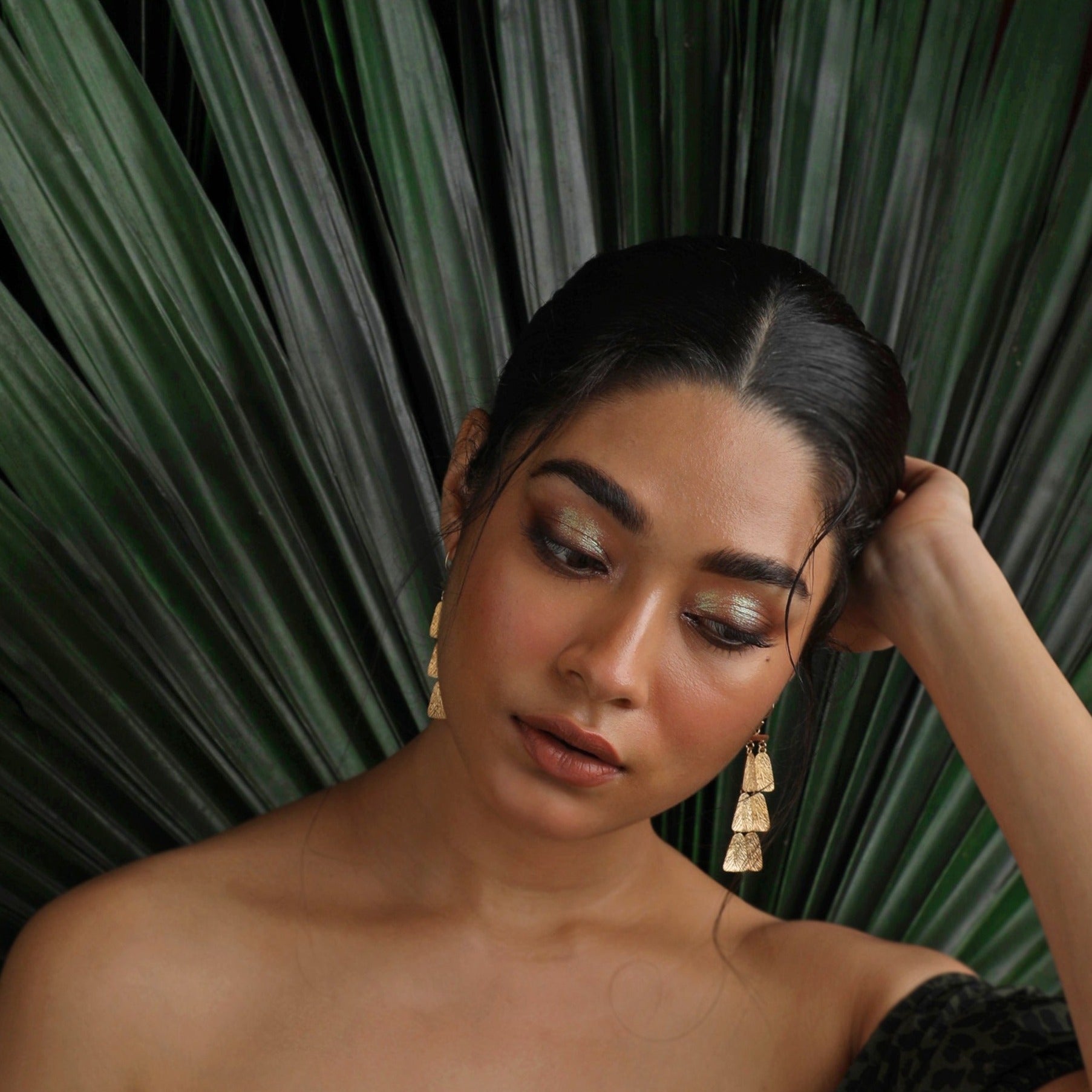 Front view of woman wearing pair of gold dangle earrings. Green fronds behind her.