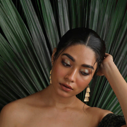 Front view of woman wearing pair of gold dangle earrings. Green fronds behind her.