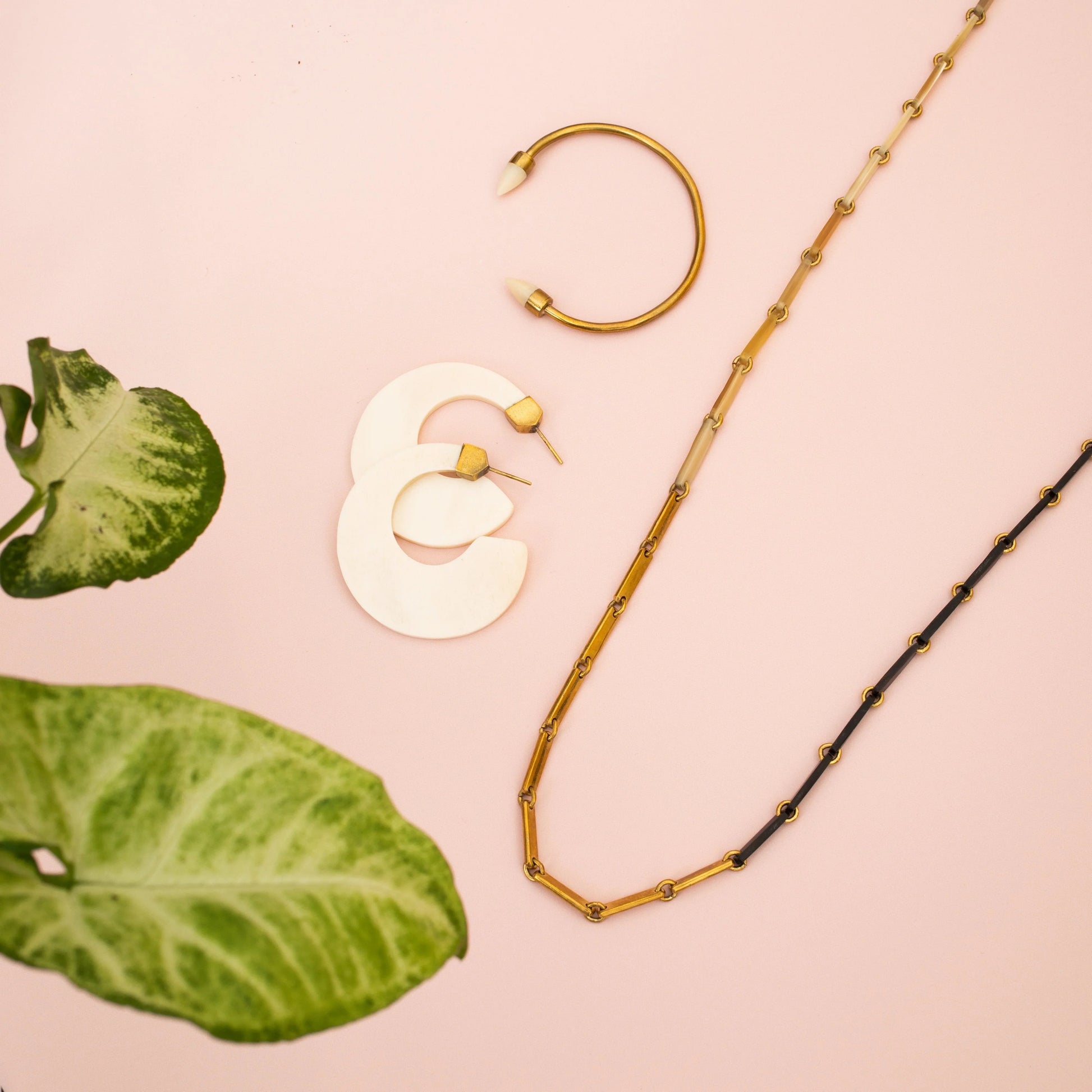 White, gold, and black link necklace, white hoop earrings, and gold bracelet on a pink background with tropical leaves.