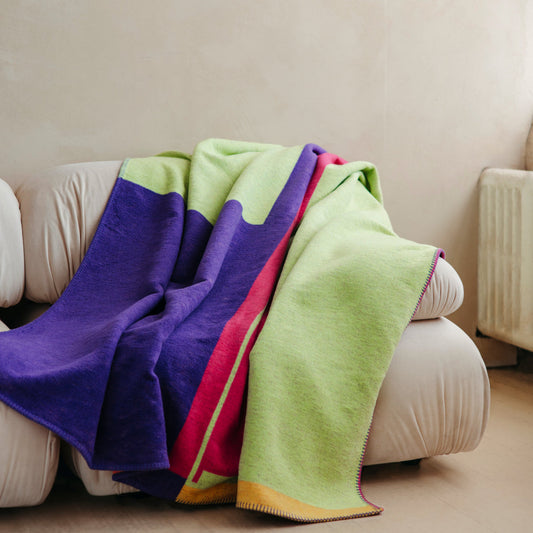 Blanket in bright purple, green, pink, and orange asymmetrical designs, draped over sofa.