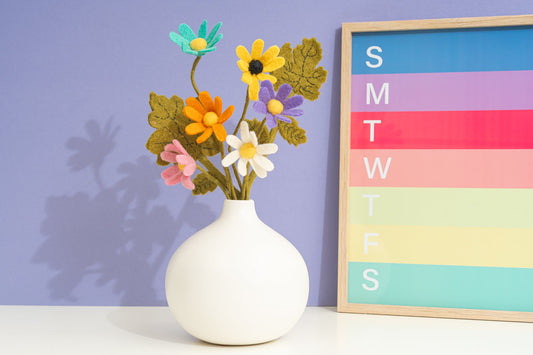 White vase with colorful handful felted flowers against a purple wall with a calendar, all fair trade | Global Goods Partners