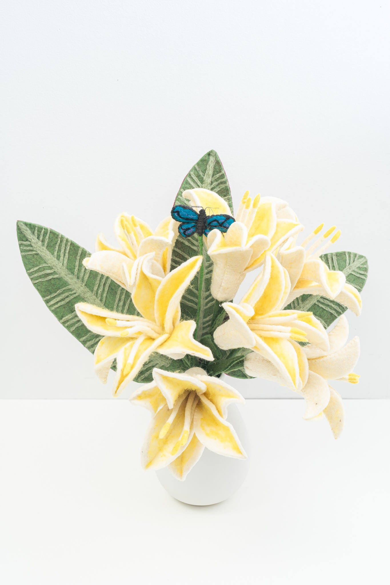 Decorative arrangement of handmade yellow felt flowers and green leaves with a blue butterfly on a white background. Made by women artisans in Nepal | Global Goods Partners