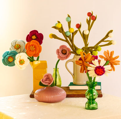 Bouquet of felt poppy flowers in magenta, blue, pink, white, orange, and light blue in a yellow vase on a table with other vases of various felt flowers.