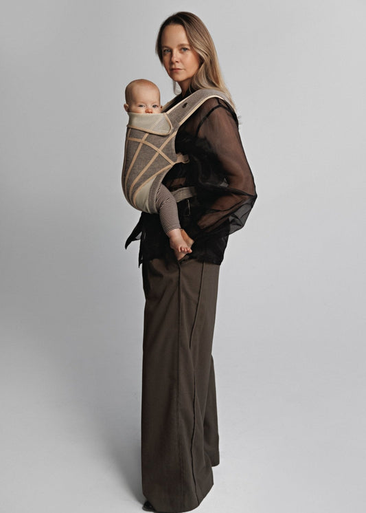 Side left view of Woman carrying baby on chest in a baby carrier. Woman is wearing black shirt and brown pants.