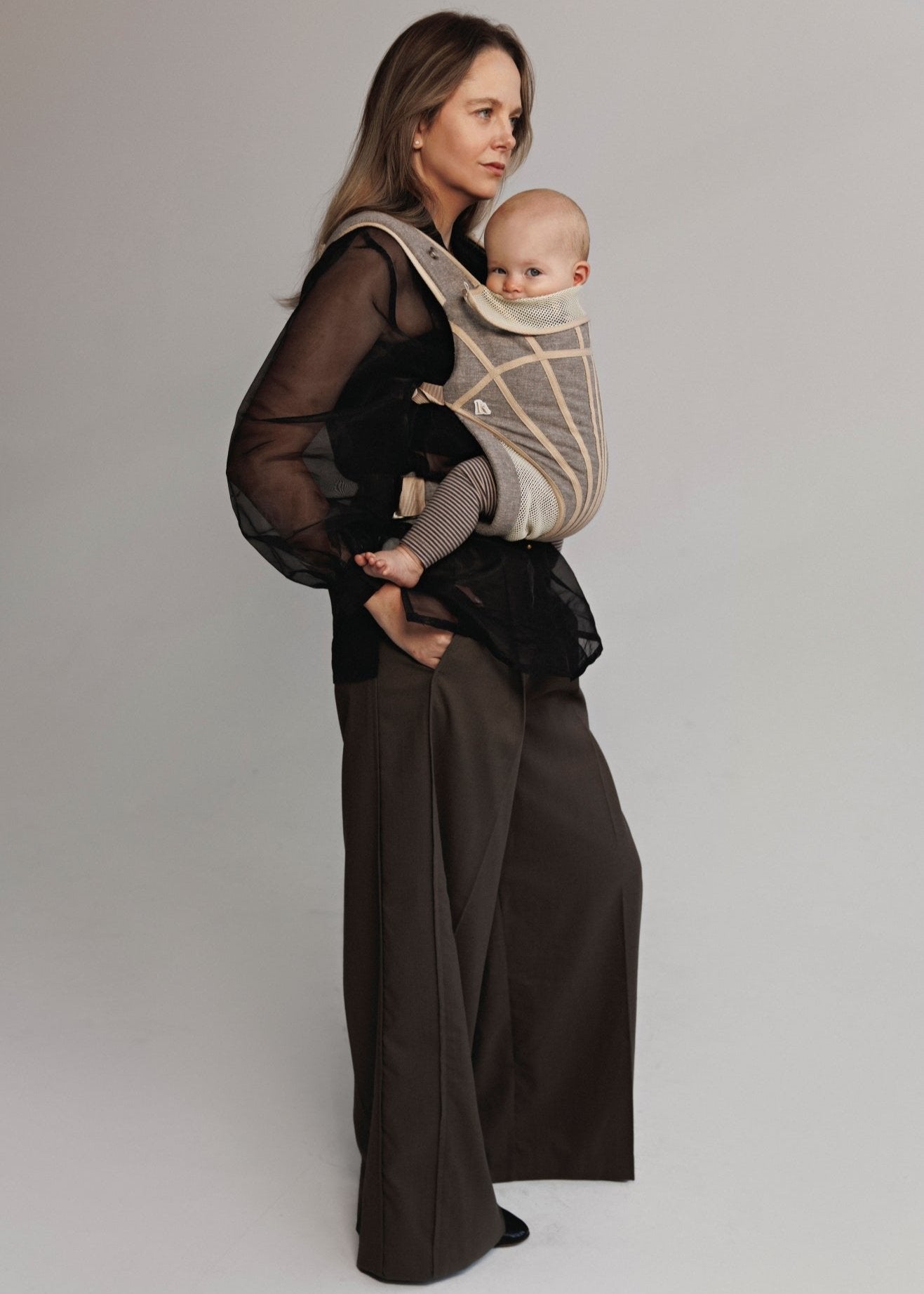 Front right view of Woman carrying baby on chest in a baby carrier. Woman is wearing black shirt and brown pants.