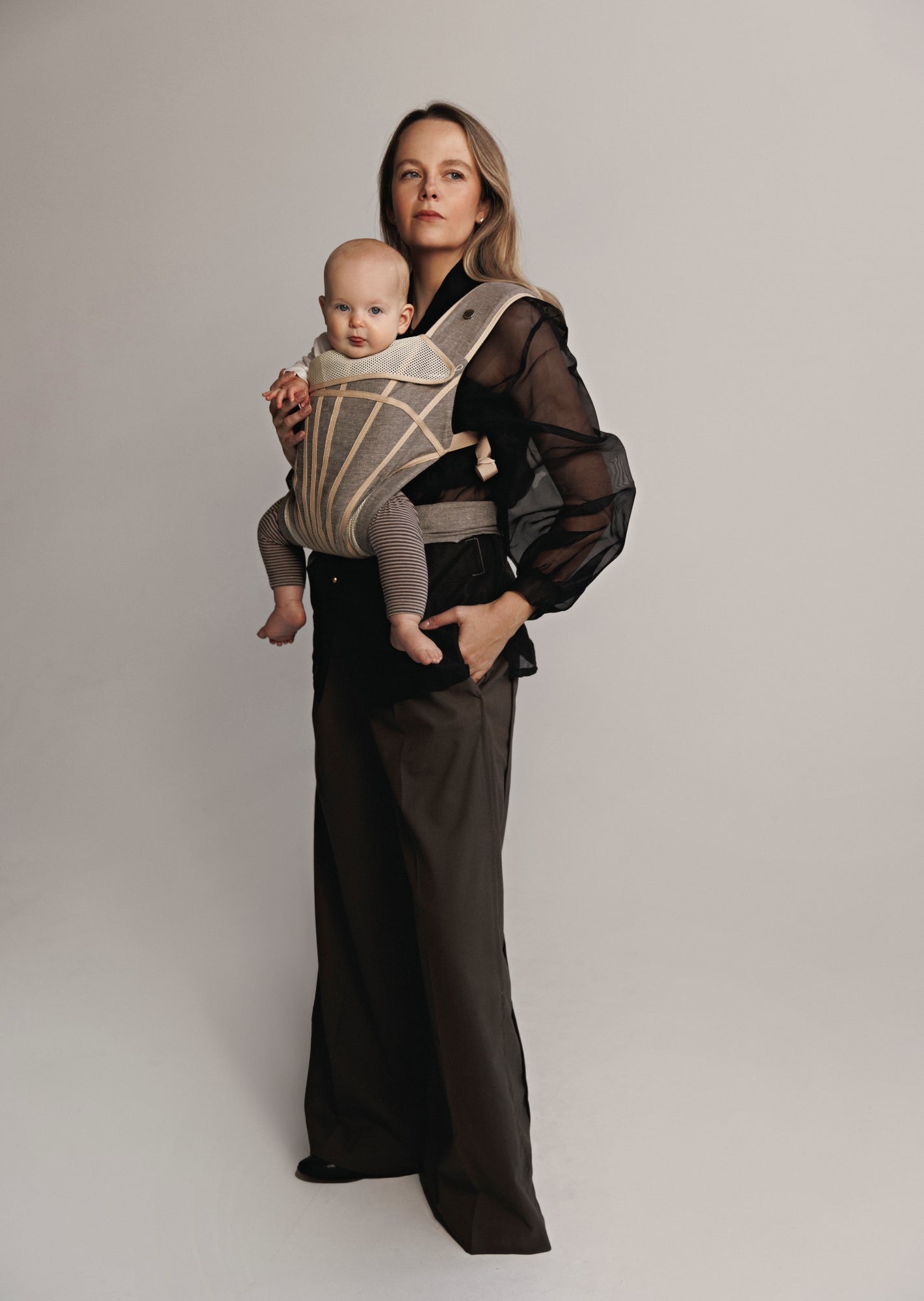 Left front view of Woman carrying baby on chest in a baby carrier. Woman is wearing black shirt and brown pants.