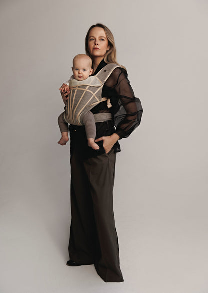 Left front view of Woman carrying baby on chest in a baby carrier. Woman is wearing black shirt and brown pants.