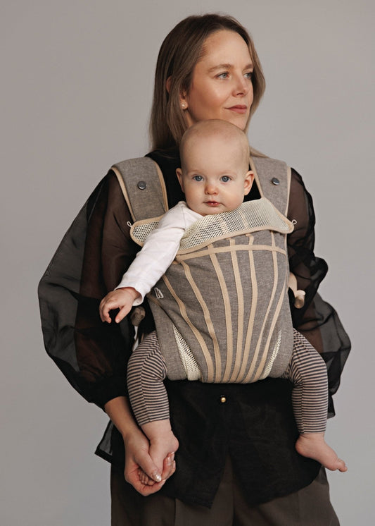 Woman carrying baby on chest in a baby carrier that is grey with stripes. Woman is wearing black shirt and brown pants.