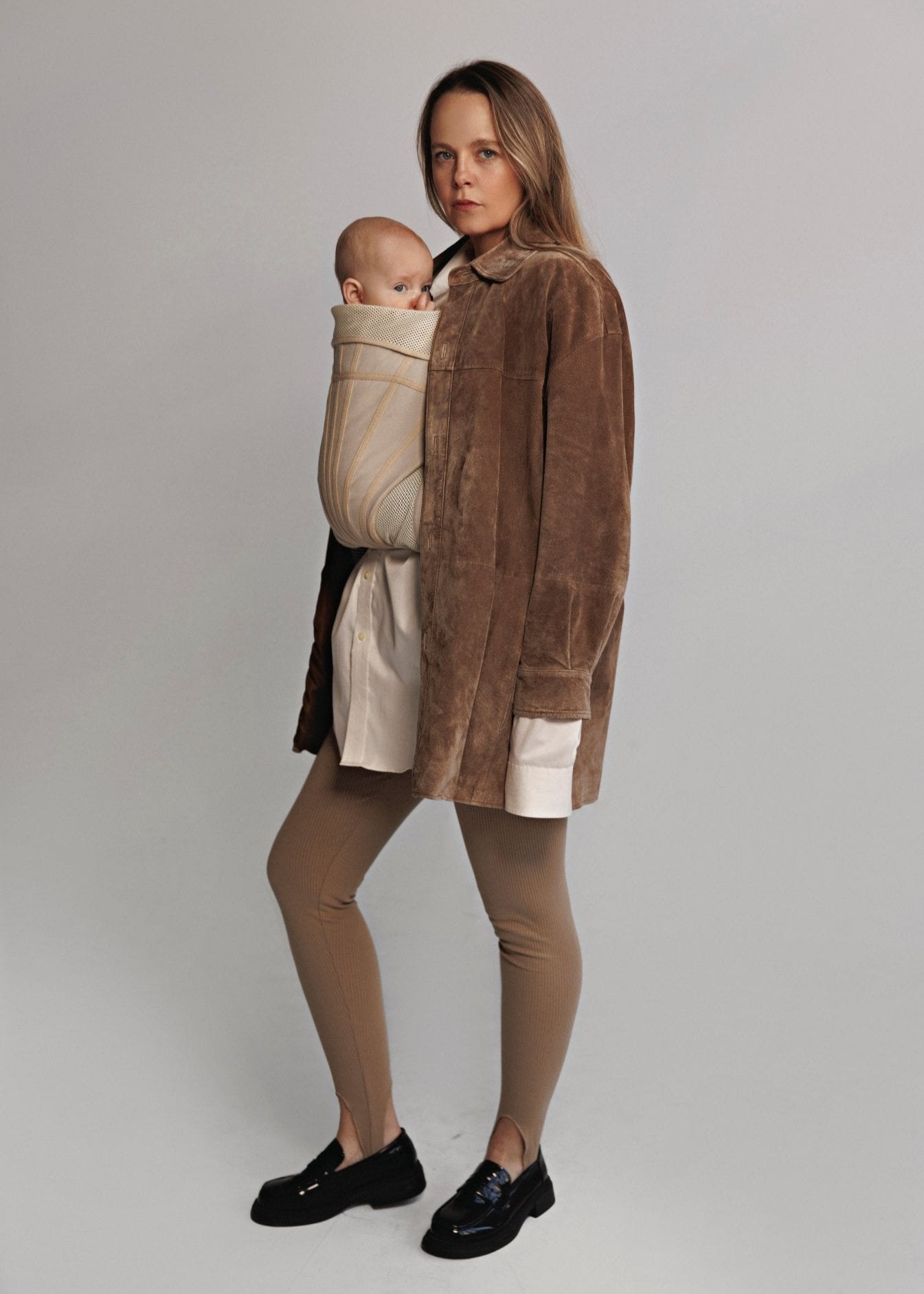 Front side view of Woman carrying baby on chest in a baby carrier. Woman is wearing brown sweater, white long shirt, brown tights, and black shoes.