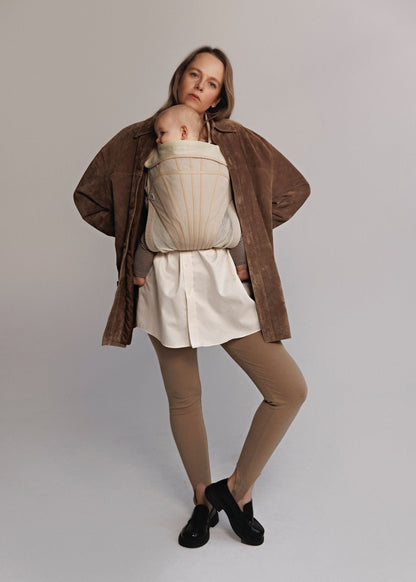 Woman carrying baby on chest in a baby carrier. Woman is wearing brown sweater, white long shirt, brown tights, and black shoes.