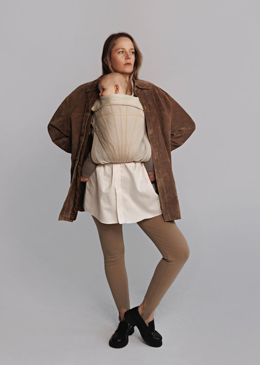 Woman carrying baby on chest in a baby carrier. Woman is wearing brown sweater, white long shirt, brown tights, and black shoes.