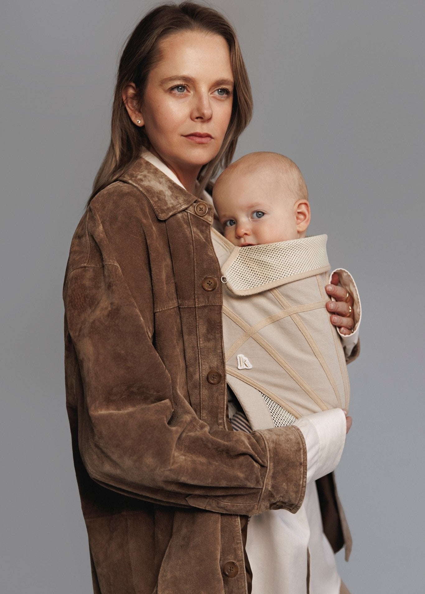 Right front side view of woman carrying baby on chest in a baby carrier. Woman is weariing white long shirt and brown sweater..