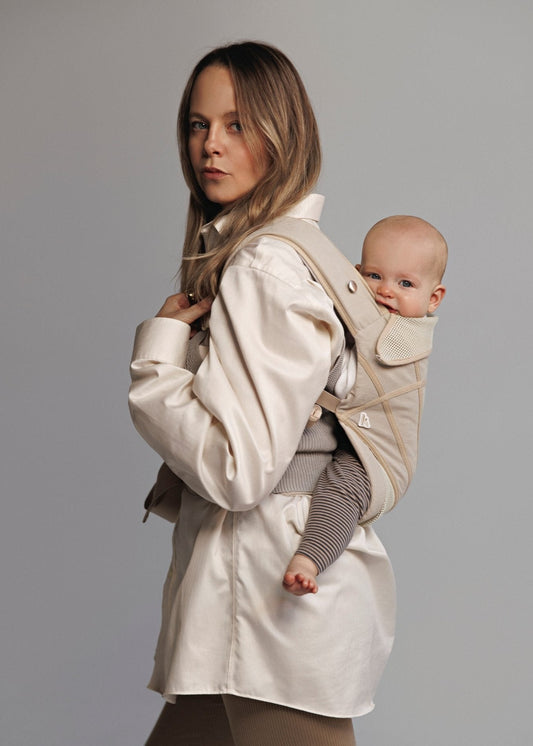 Front left partial view of woman carrying baby on back in a baby carrier. Woman is weariing white long shirt.