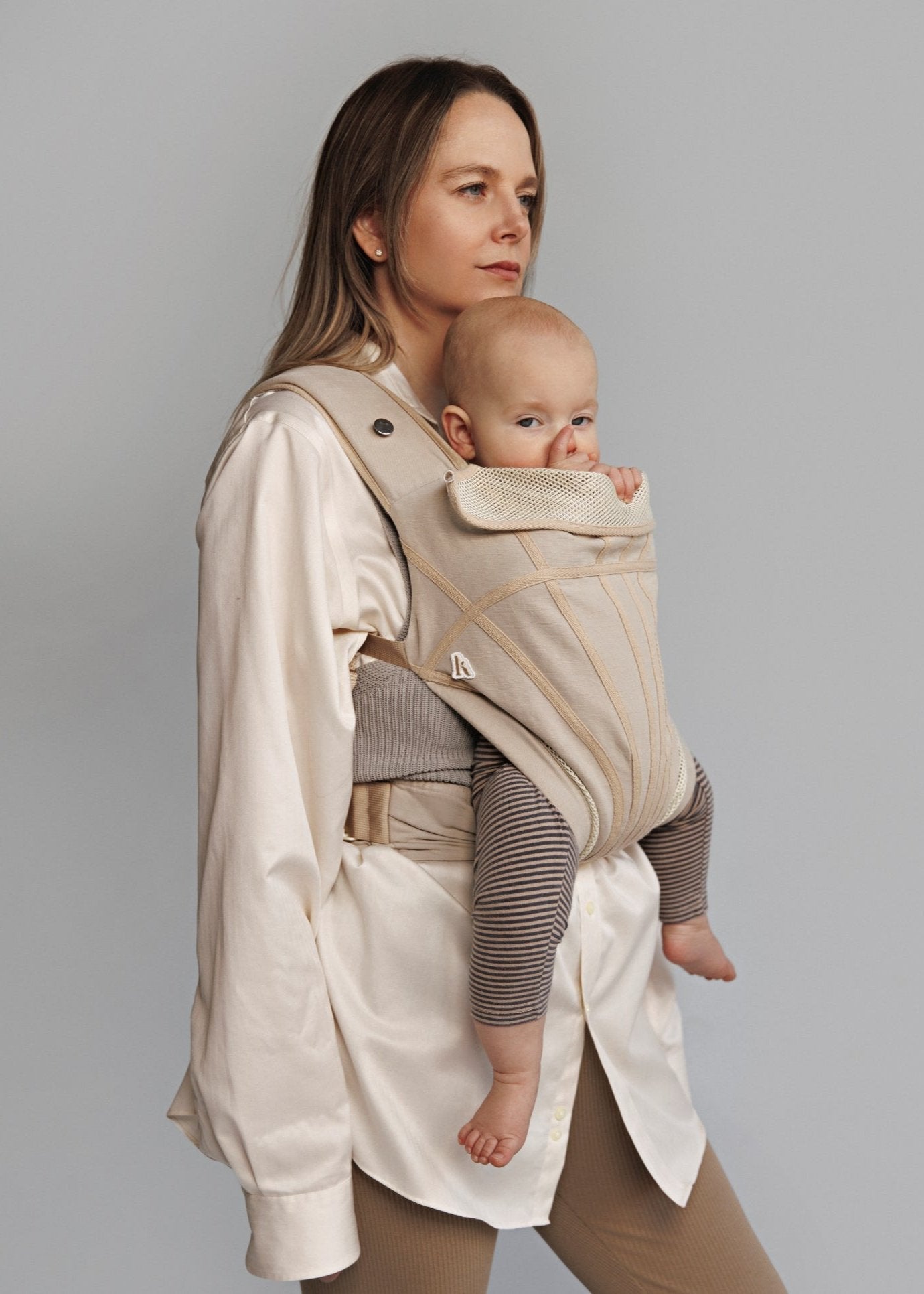 Partial front view of woman carrying baby on her front in a baby carrier. Woman is wearing white long shirt.