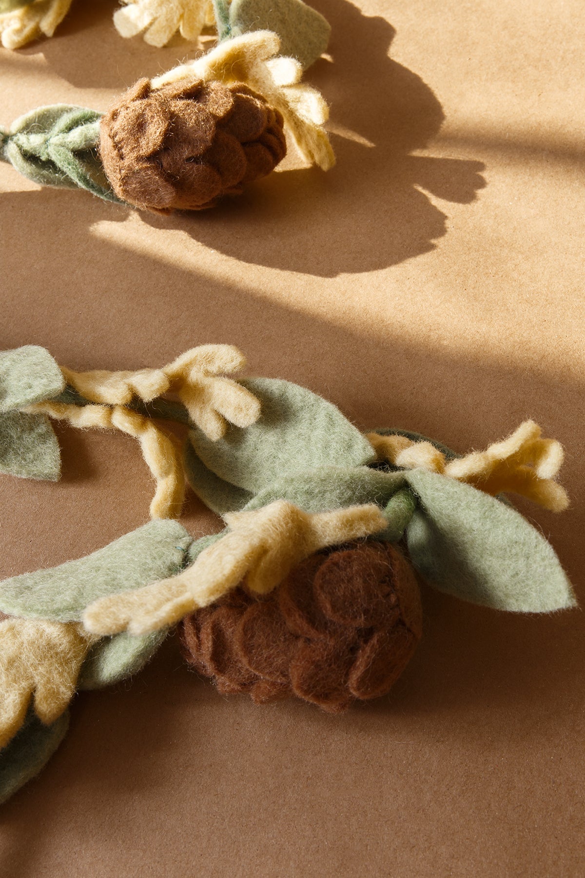 Close up of felt garland with green leaves and brown pinecones laying on tan table.