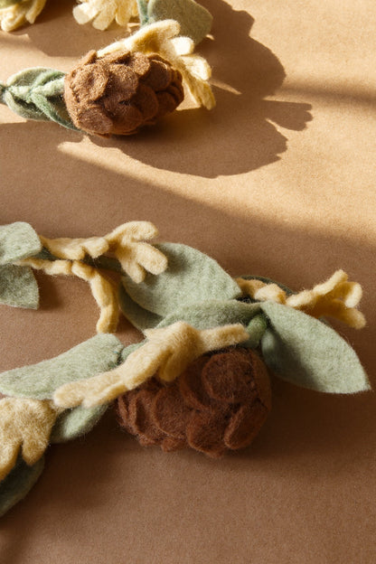 Close up of felt garland with green leaves and brown pinecones laying on tan table.