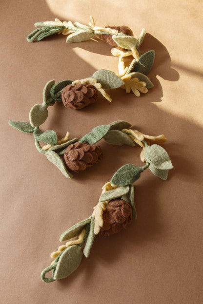 Felt garland with green leaves and brown pinecones laying on tan table.