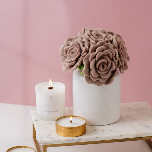 Pink rose felt floral arrangement in a white vase, sitting on a table with two candles.