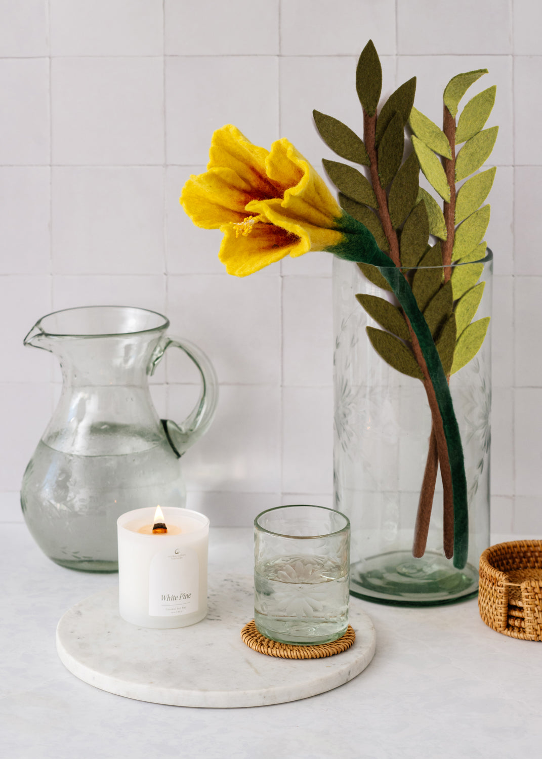 Etched clear glass vase with felt leaves and yellow flower next to pitcher and candle.