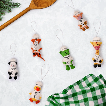 Scattered felt ornaments of animals in Chef's hats and coats showing a panda, duck, giraffe, frog, pig, and tiger on table with cooking spoon and green and white checkered kitchen towel.