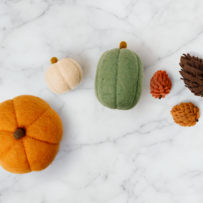 Three felt pumpkins, one large orange pumpkin, one medium oblong green one, and one small tan one spread out on white counter with pinecones.