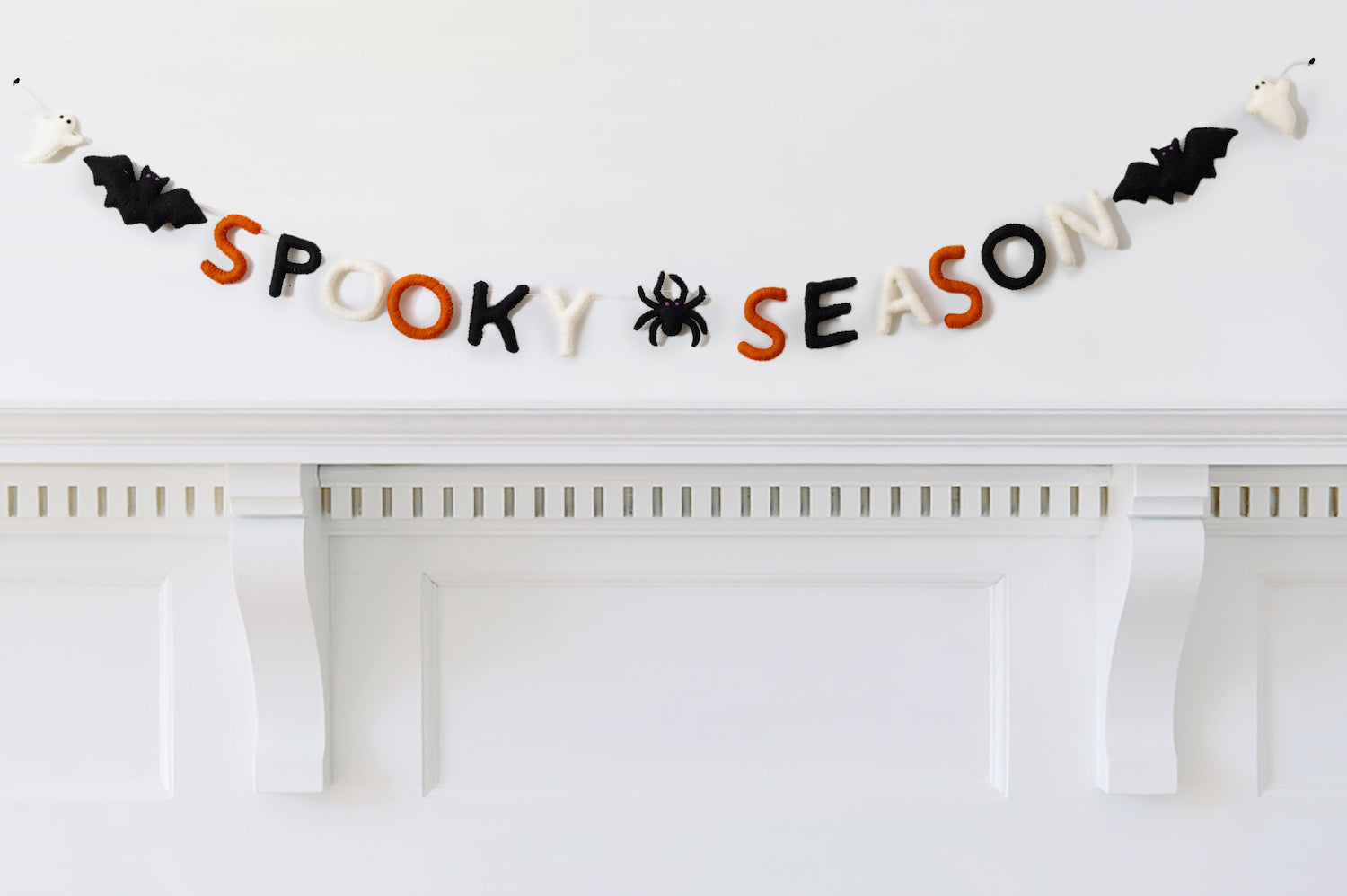 Felt garland that says "Spooky Season" in orange, black, and white letters with a black spider between the words and a bat and ghost on the ends hanging over white mantel.