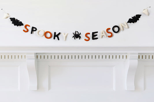 Felt garland that says "Spooky Season" in orange, black, and white letters with a black spider between the words and a bat and ghost on the ends hanging over white mantel.