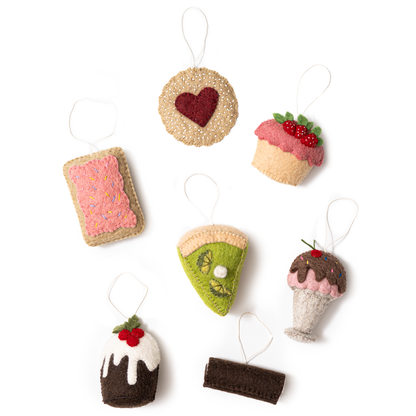 Scattered felt food ornaments of a pink pop tart, a Linzer cookie, a strawberry shortcake, key lime pie, ice cream sundae, Christmas pudding, and a Swiss roll cake.