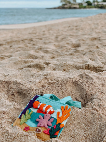 Felt bag with under the sea finger puppets of an orange and white clown fish, a pink octopus, and a grey shark laying on the beach.