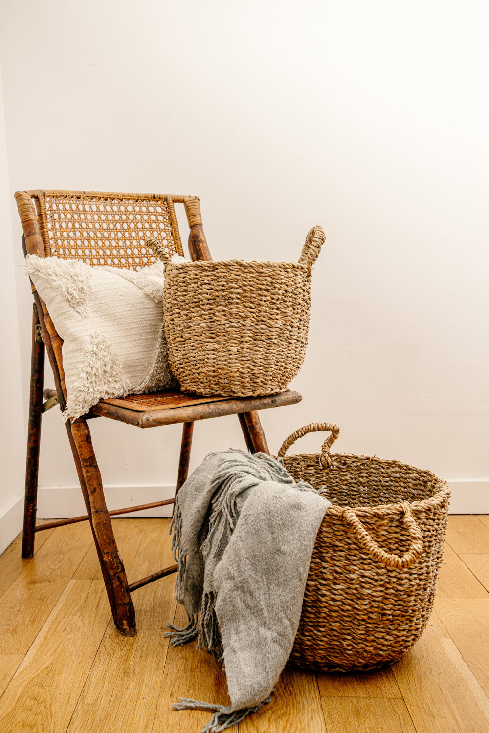 Set of two brown woven baskets, smaller on chair with pillow, larger on brown floor with blanket.