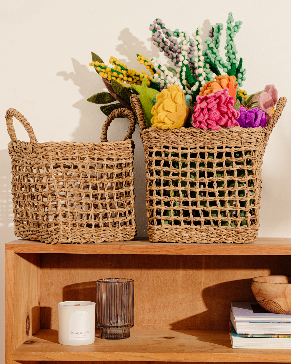 Set of two brown woven baskets with net design, one containing felt flowers, on shelf with miscellaneous items.