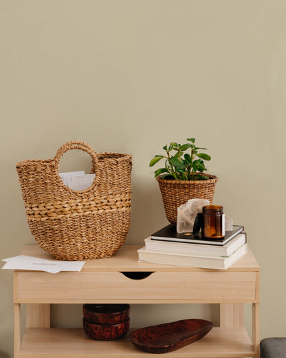 Brown woven basket with round handles on brown table with books and plant.