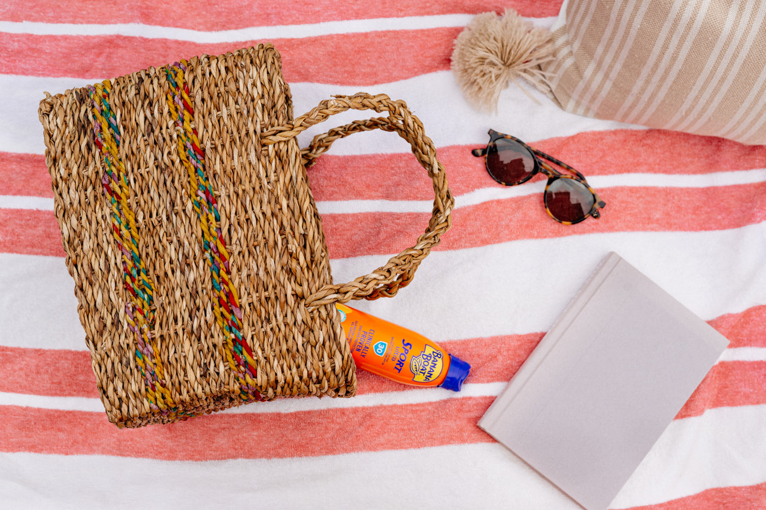 Brown woven basket laying on orange striped beach blanket, next to beach accessories.