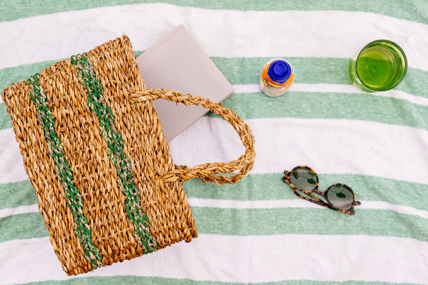 Woven brown basket with green stripes, laying on green striped towel next to sunglasses and other beach accessories.