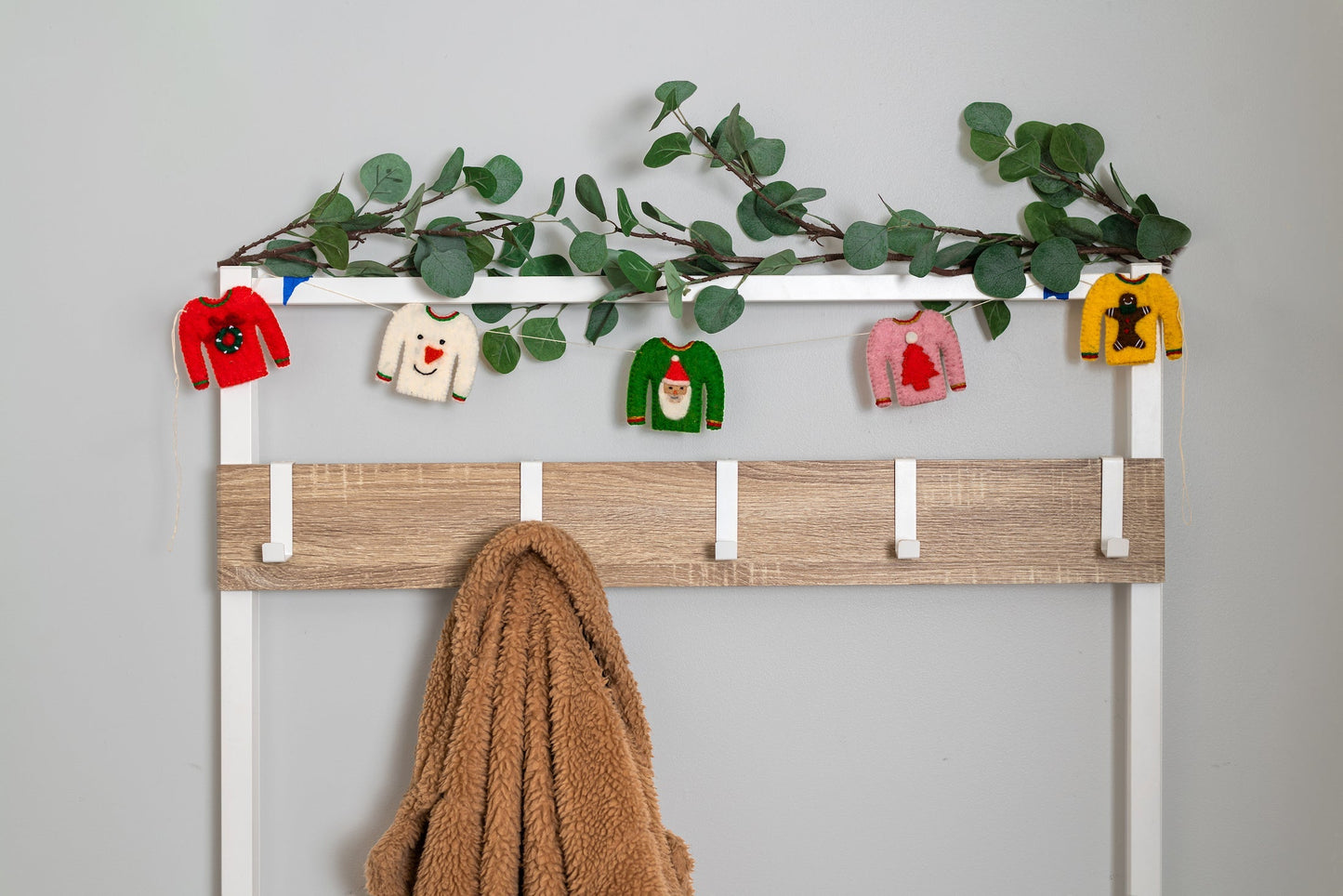 Garland of tiny holiday sweaters hanging on clothing hooks, one red with a wreath, white with snowman, pink with red tree, green with Santa, and yellow with a gingerbread man.