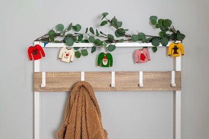Garland of tiny holiday sweaters hanging on clothing hooks, one red with a wreath, white with snowman, pink with red tree, green with Santa, and yellow with a gingerbread man.