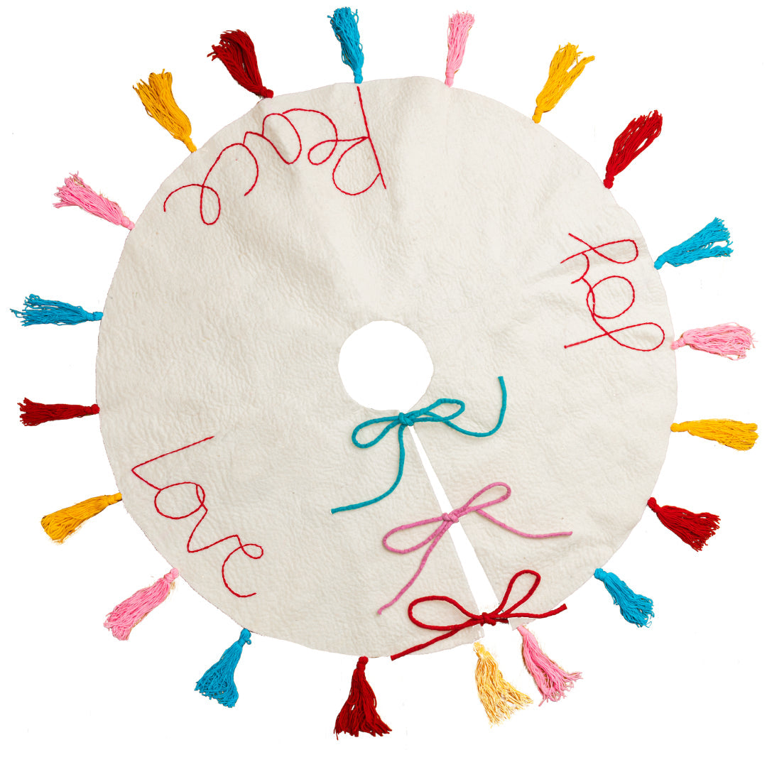 Top view of white felt Christmas tree skirt with multicolored tassels around the edges and the words "peace" "love" and 'joy' embroidered.