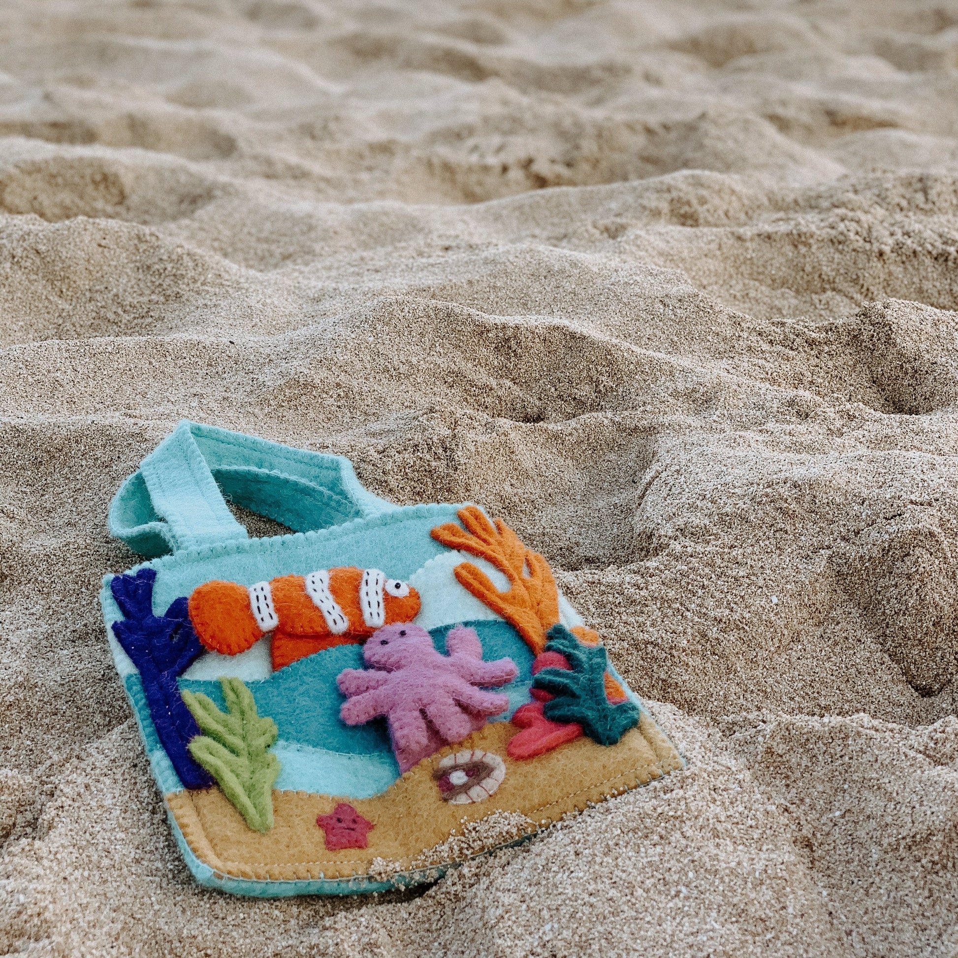 Felt bag with under the sea finger puppets of an orange and white clown fish, a pink octopus, and a grey shark laying on the sand.
