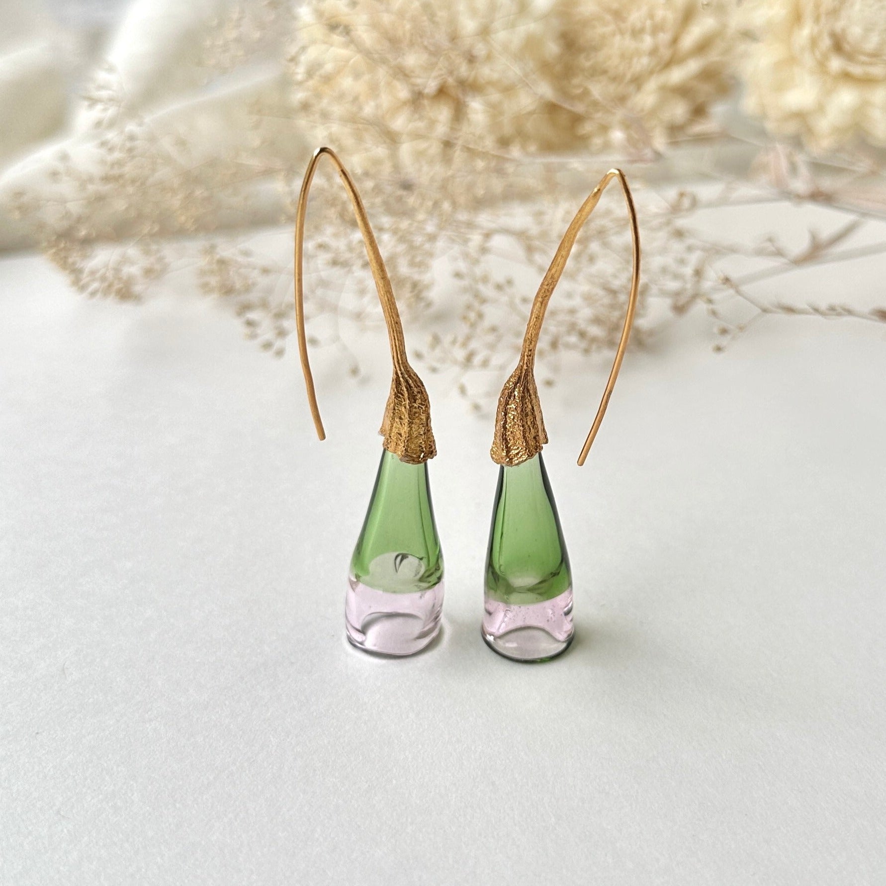 Pair of green and pale pink glass earrings with gold colored findings on grey background. Small dried flowers above image.
